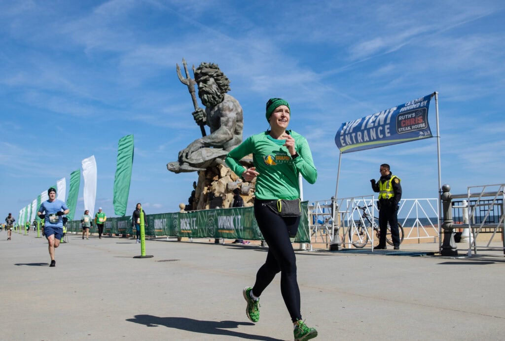 A smiling female runner in a green shirt races past a large Neptune statue during a vibrant outdoor event by the beach for Shamrock Marathon.