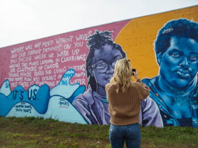 A person with long blonde hair takes a photo in front of a colorful mural in the ViBe Creative District featuring two portraits and poetic text about water and opportunity.