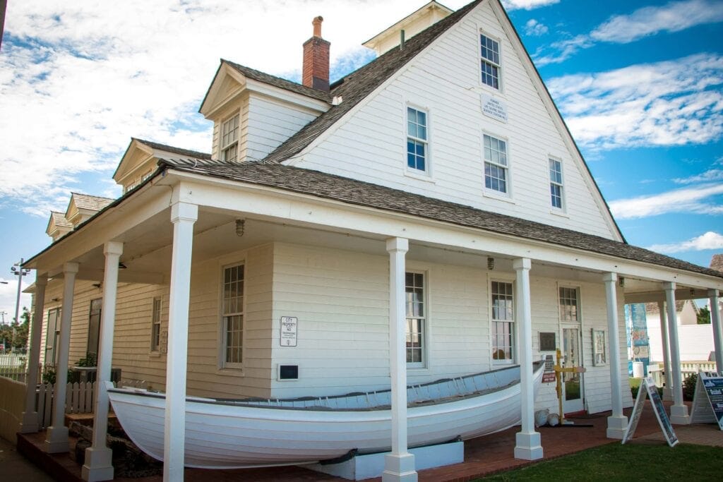The historic Surf and Rescue Museum with a porch featuring a rescue boat displayed prominently in front, under a blue sky with clouds at Virginia Beach Boardwalk.