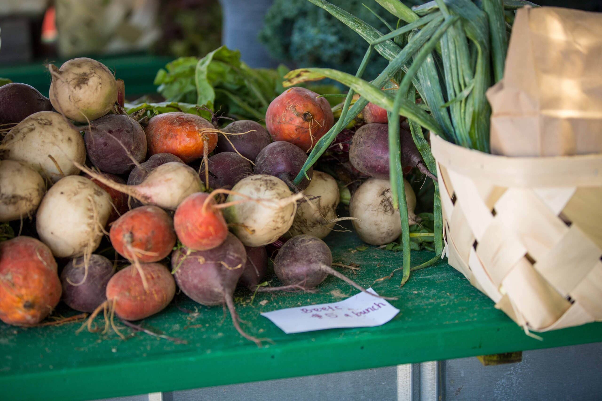 A colorful array of various radishes, including purple and white, displayed at Old Beach Farmer's Market alongside green onions and a brown basket.