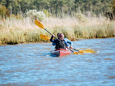 A woman and man paddle a kayak through the calm waters with lush tall grass and woods behind them in False Cape State Park.