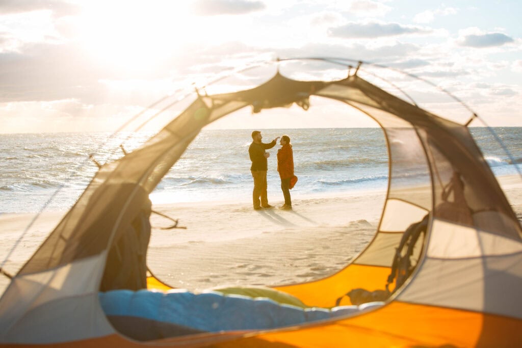 A couple stands on the beach in False Cape State Park, framed by a camping tent, with the sun rising over the ocean in the background.