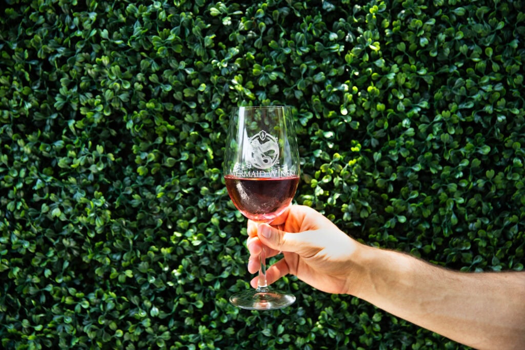 A hand holding a glass of red wine with a logo of Mermaid Winery, set against a backdrop of lush green foliage on a sunny spring day.