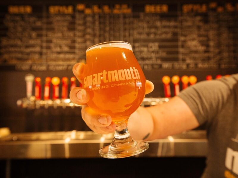 A hand holding a glass of amber beer with the logo of Smartmouth Brewing Company, set against a menu board and taps in a brewery.