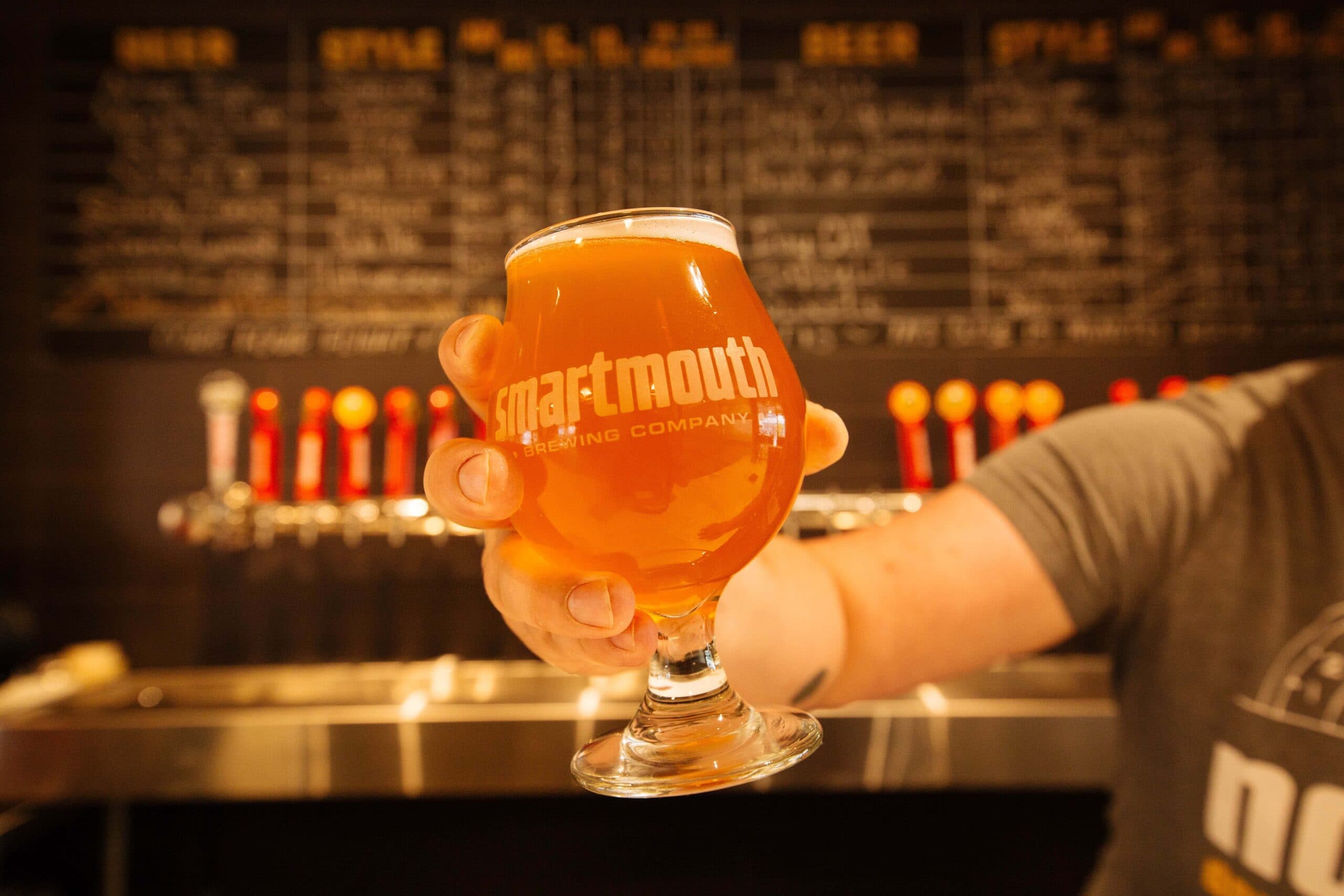 A hand holding a glass of amber beer with the logo of Smartmouth Brewing Company, set against a menu board and taps in a brewery.