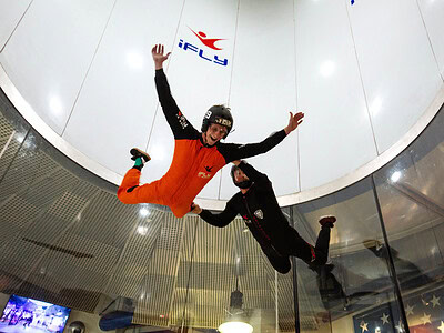 Two excited people doing aerial skydiving maneuvers in a wind tunnel with partial glass windows at iFly Virginia Beach