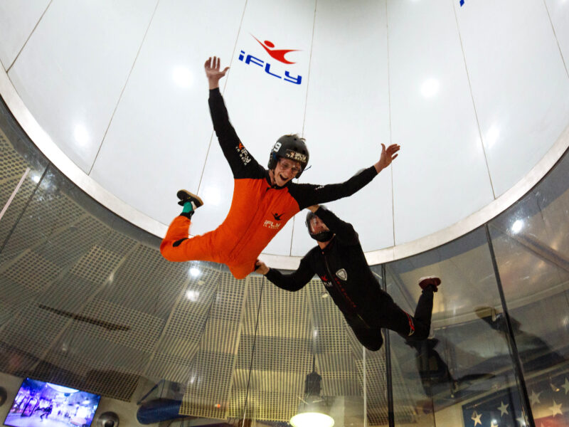 Two excited people doing aerial skydiving maneuvers in a wind tunnel with partial glass windows at iFly Virginia Beach