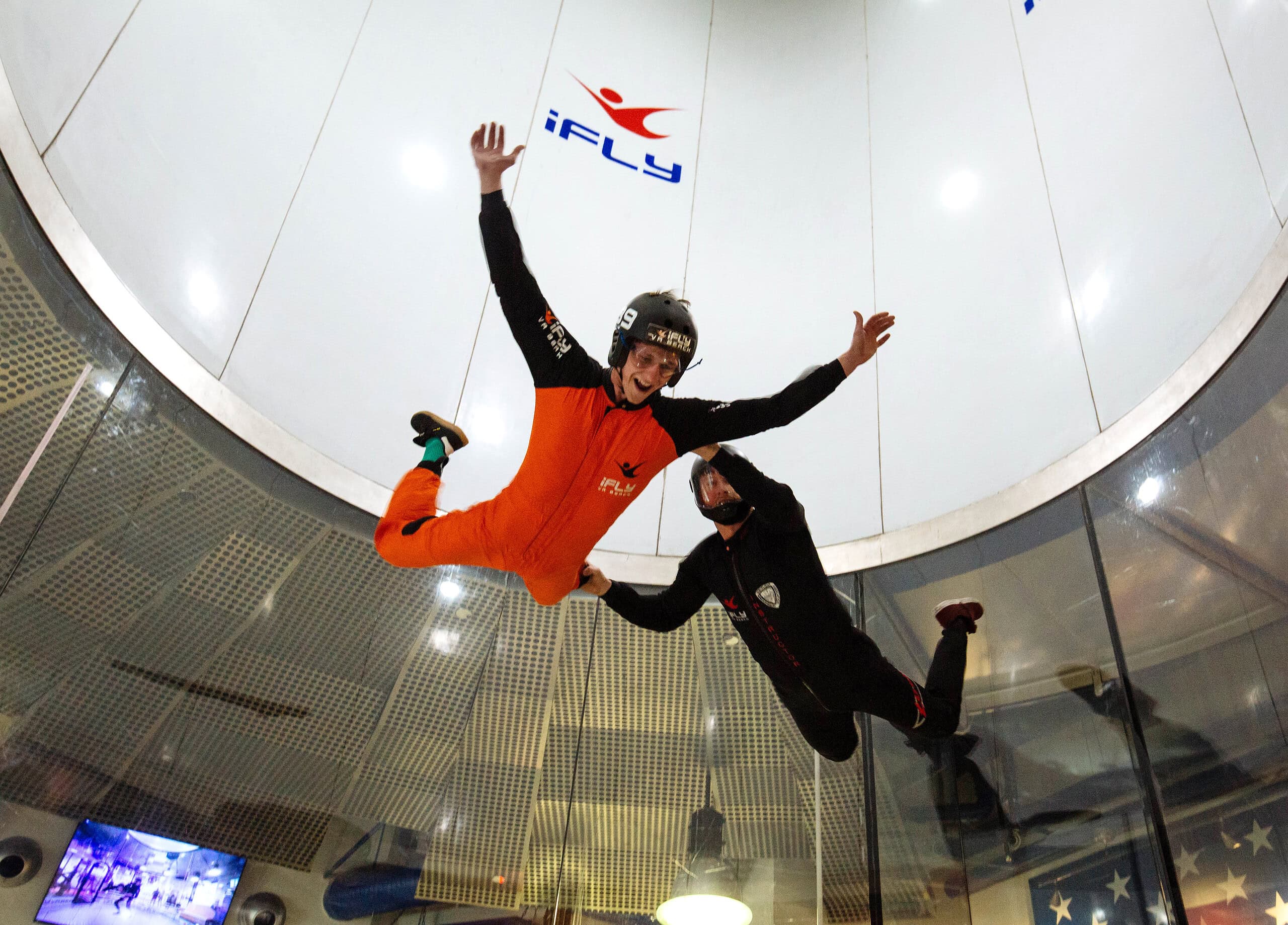 Two excited people doing aerial skydiving maneuvers in a wind tunnel with partial glass windows at iFly Virginia Beach