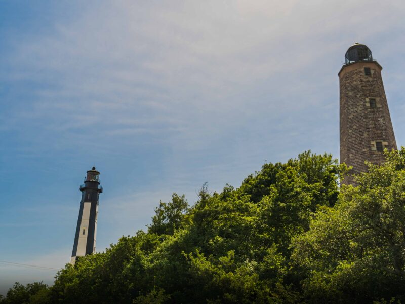 The old and the new Cape Henry lighthouses stand on a lush green hillside under a clear blue sky, showcasing distinct architectural styles.