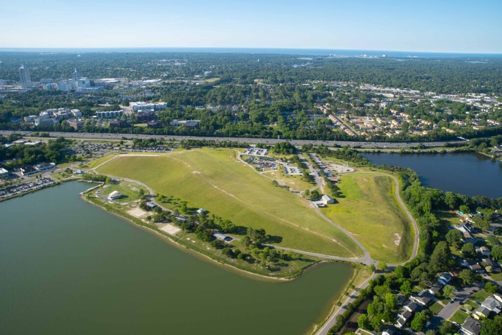 Aerial view of Mount Trashmore Park with two bodies of water, suburban neighborhoods, and highway surrounding it w/a view of the coastline & Town Center in the background