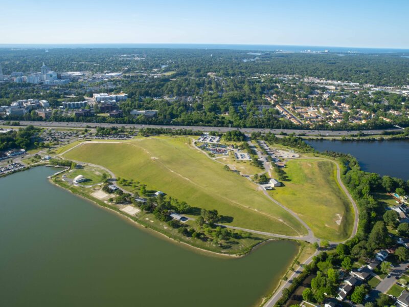 Aerial view of Mount Trashmore Park with two bodies of water, suburban neighborhoods, and highway surrounding it w/a view of the coastline & Town Center in the background