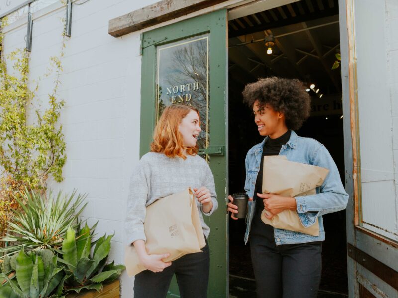 Two excited women standing and talking outside North End Bag Company with wrapped items from the store.