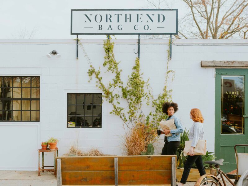 Two women with bags in their arms walking past North End Bag Co, a white industrial building with foliage growing up the side.