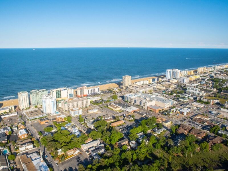 Aerial view of a coastal city with sandy beaches, high-rise buildings, and a clear blue ocean under a bright sky at Virginia Beach Oceanfront.