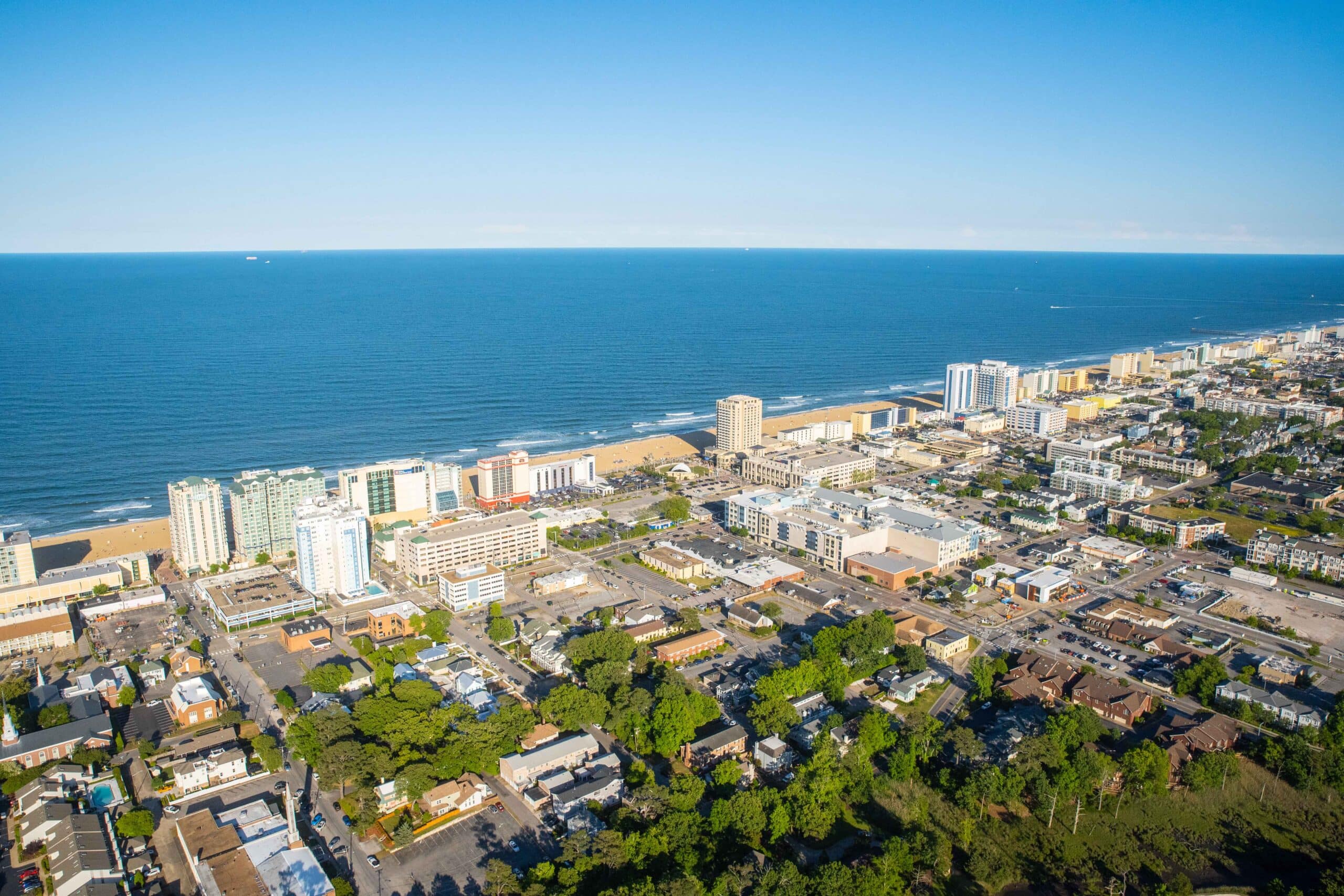 Aerial view of a coastal city with sandy beaches, high-rise buildings, and a clear blue ocean under a bright sky at Virginia Beach Oceanfront.