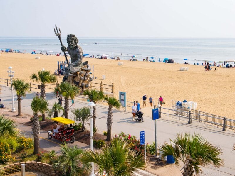 A vibrant beach scene at Virginia Beach Oceanfront featuring a large statue of King Neptune, palm trees, pedestrians, and colorful umbrellas along the sandy shore.
