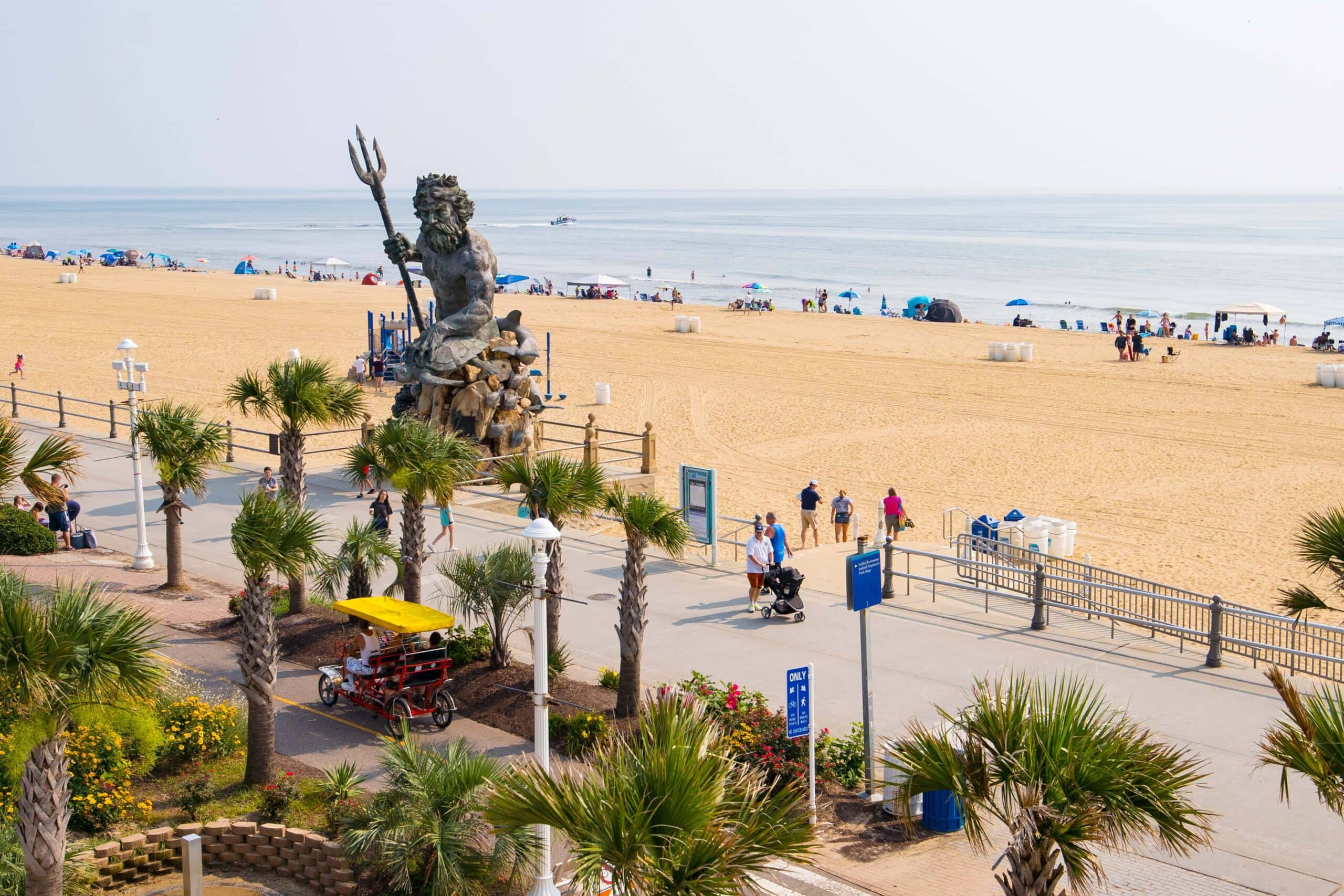 A vibrant beach scene at Virginia Beach Oceanfront featuring a large statue of King Neptune, palm trees, pedestrians, and colorful umbrellas along the sandy shore.