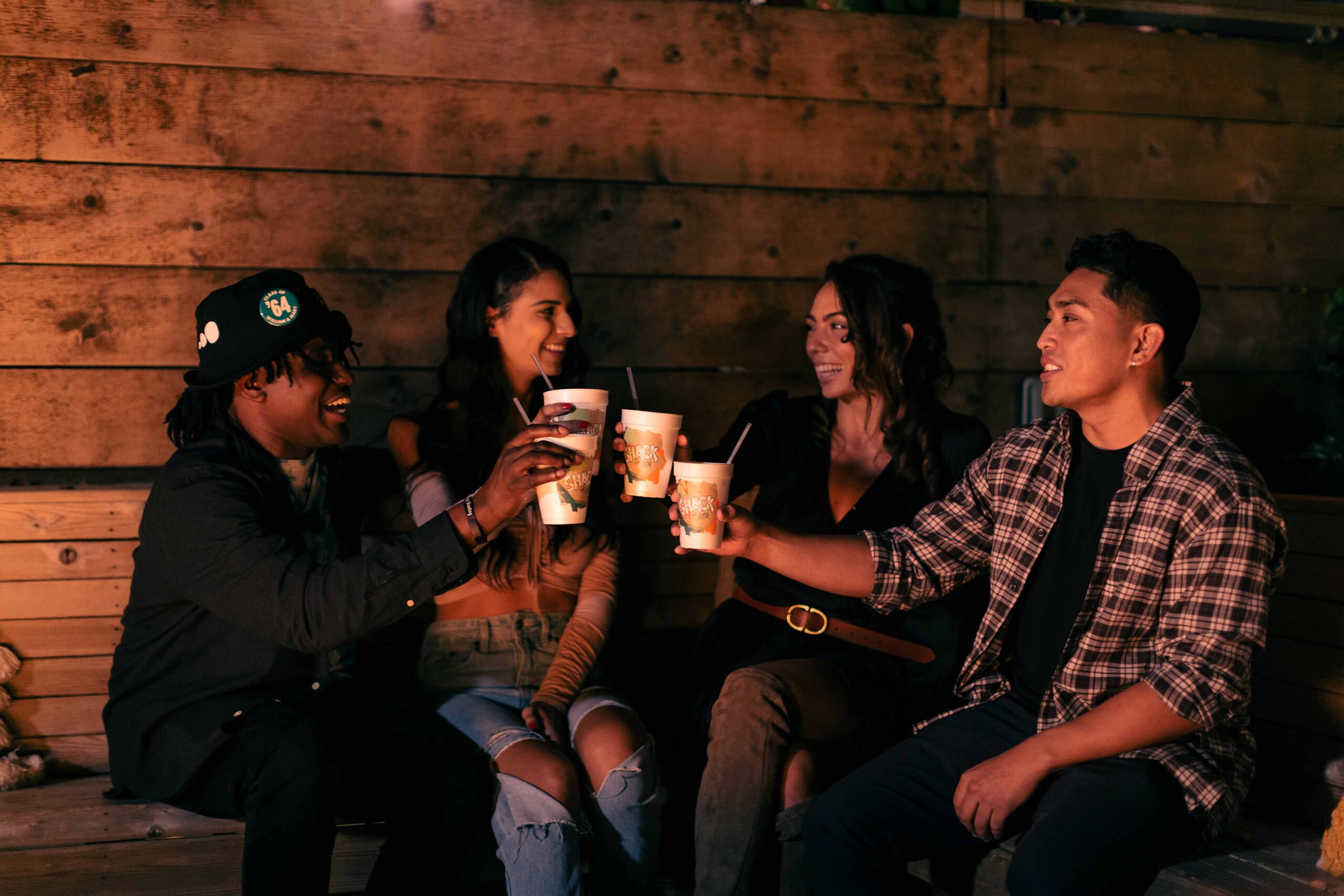 A group of four friends enjoys drinks together, celebrating and laughing in a cozy, dimly-lit outdoor setting at The Shack in Virginia Beach.