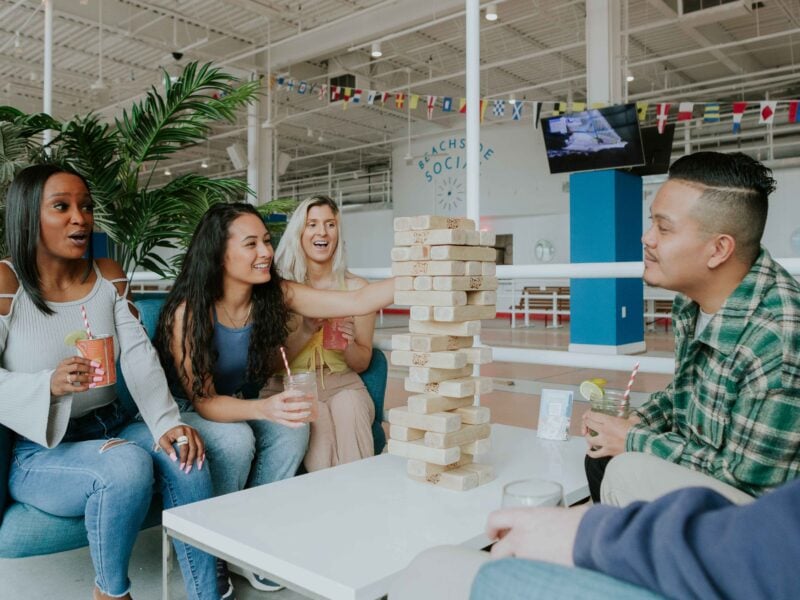A group of friends enjoys drinks while playing Jenga in a lively social setting, surrounded by greenery and vibrant decor at Beachside Social.