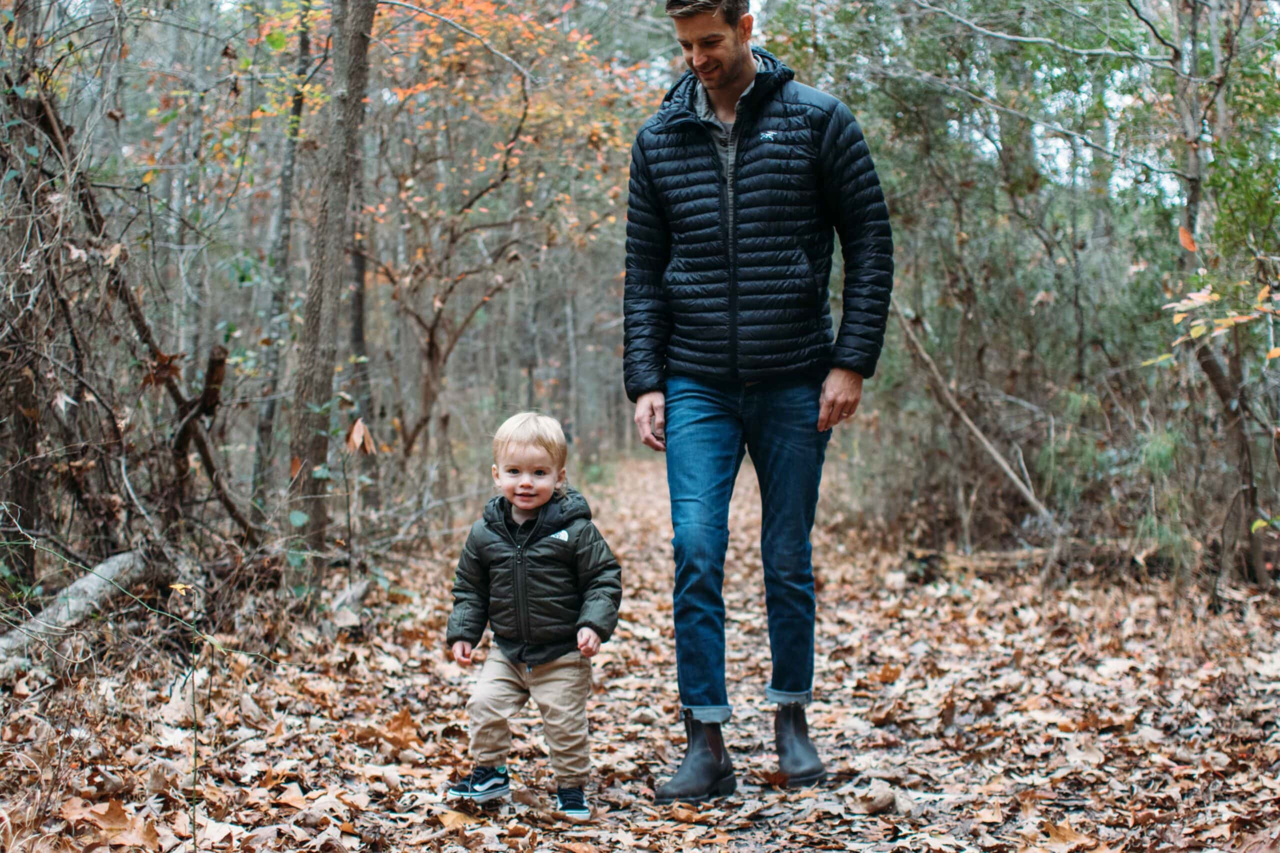 A father and son walk together on a leaf-covered path in a forest, surrounded by trees with winter foliage in First Landing State Park.