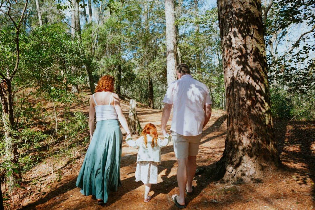 A family walks hand-in-hand along a wooded trail in First Landing State park, with a woman in a flowing skirt, a man in a light shirt, and a girl in a white dress.