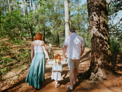A family walks hand-in-hand along a wooded trail in First Landing State park, with a woman in a flowing skirt, a man in a light shirt, and a girl in a white dress.