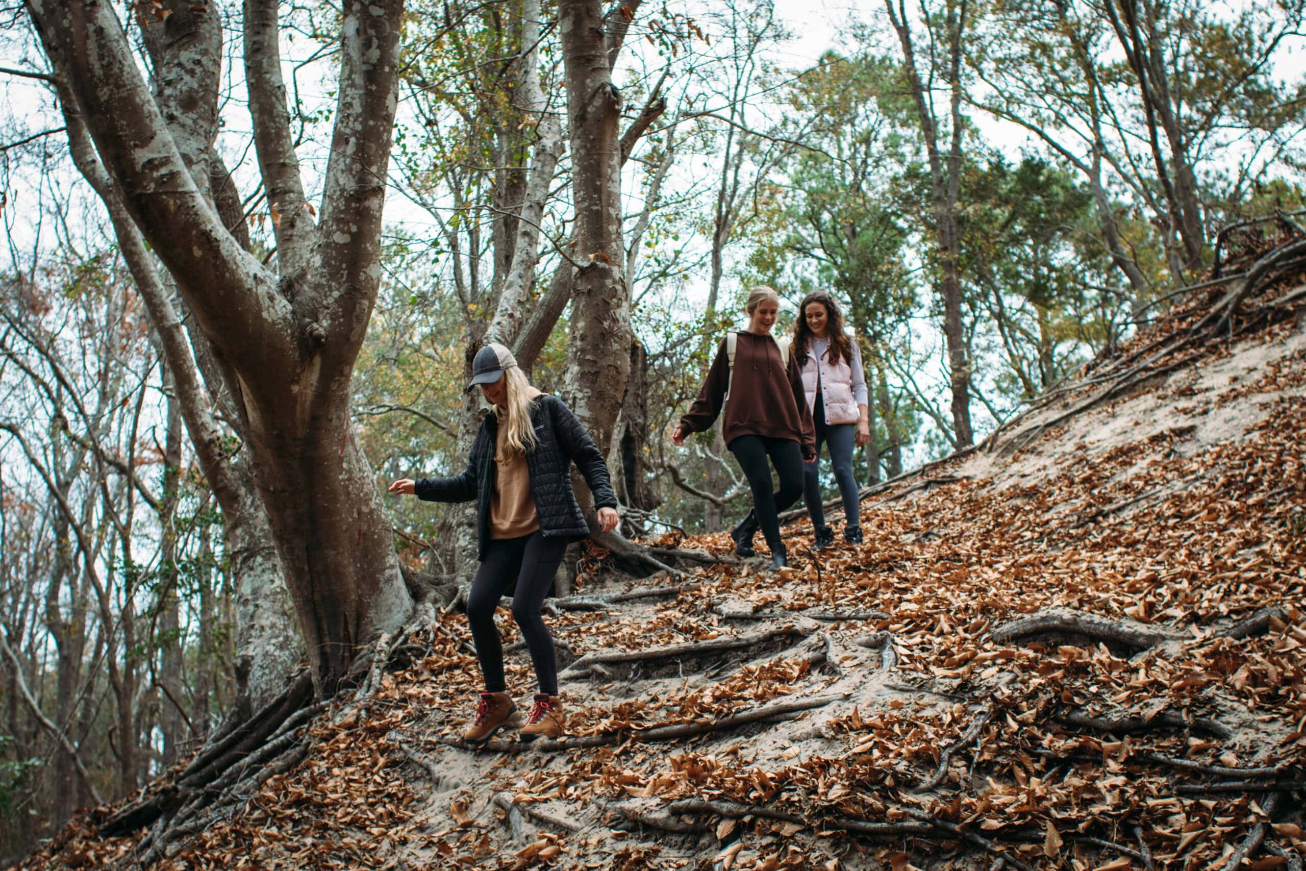 Three women navigate a leaf-covered slope in a forest, showcasing an autumn landscape with trees and earthy tones in First Landing State Park.