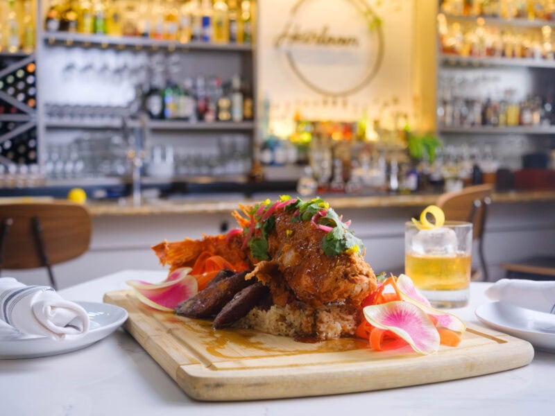 An elegantly presented dish featuring crispy whole fish garnished with plantains, herbs, and flowers, served on a wooden board with rice and a cocktail in a stylish bar setting.