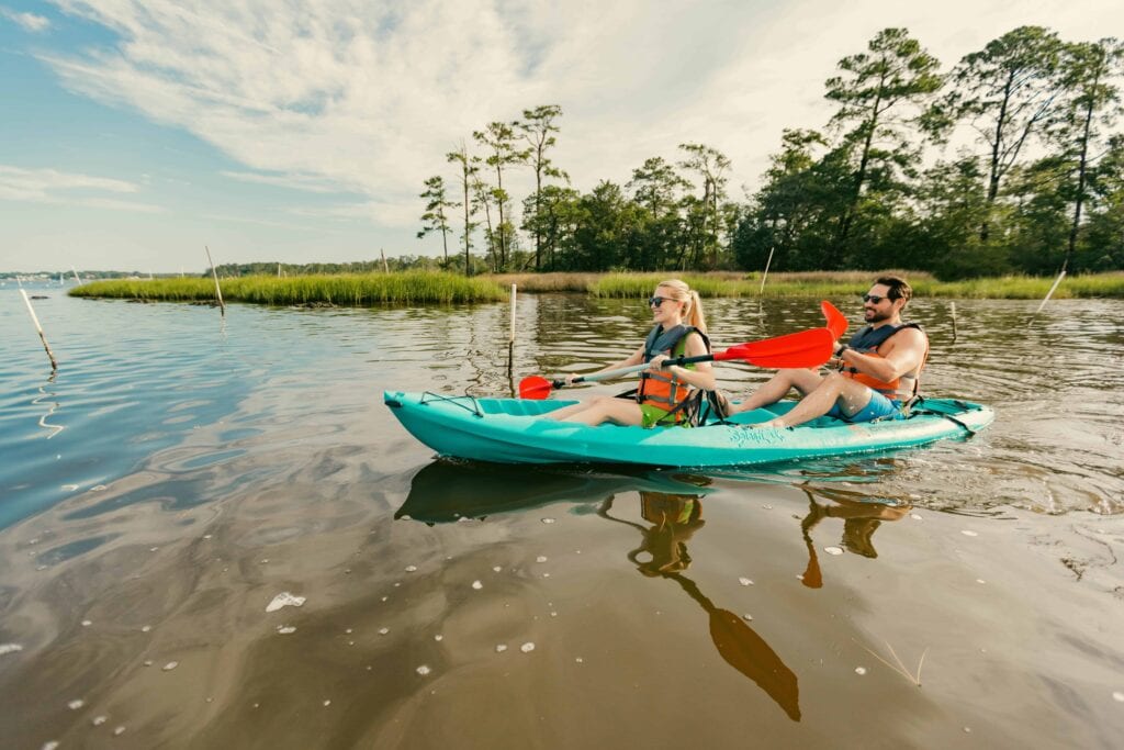 A couple kayaks in First Landing State park on a calm body of water surrounded by greenery, enjoying a sunny day with their paddles and wearing life jackets.