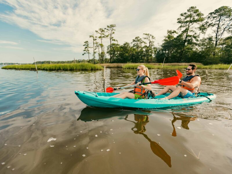 A couple kayaks in First Landing State park on a calm body of water surrounded by greenery, enjoying a sunny day with their paddles and wearing life jackets.