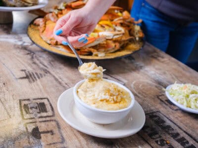 Une main tenant une cuillerée de soupe crémeuse de crabe femelle au-dessus d’un bol sur une table en bois, avec un plateau de crabe en arrière-plan et du coleslaw en accompagnement, chez Margie and Ray's Crabhouse & Restaurant.