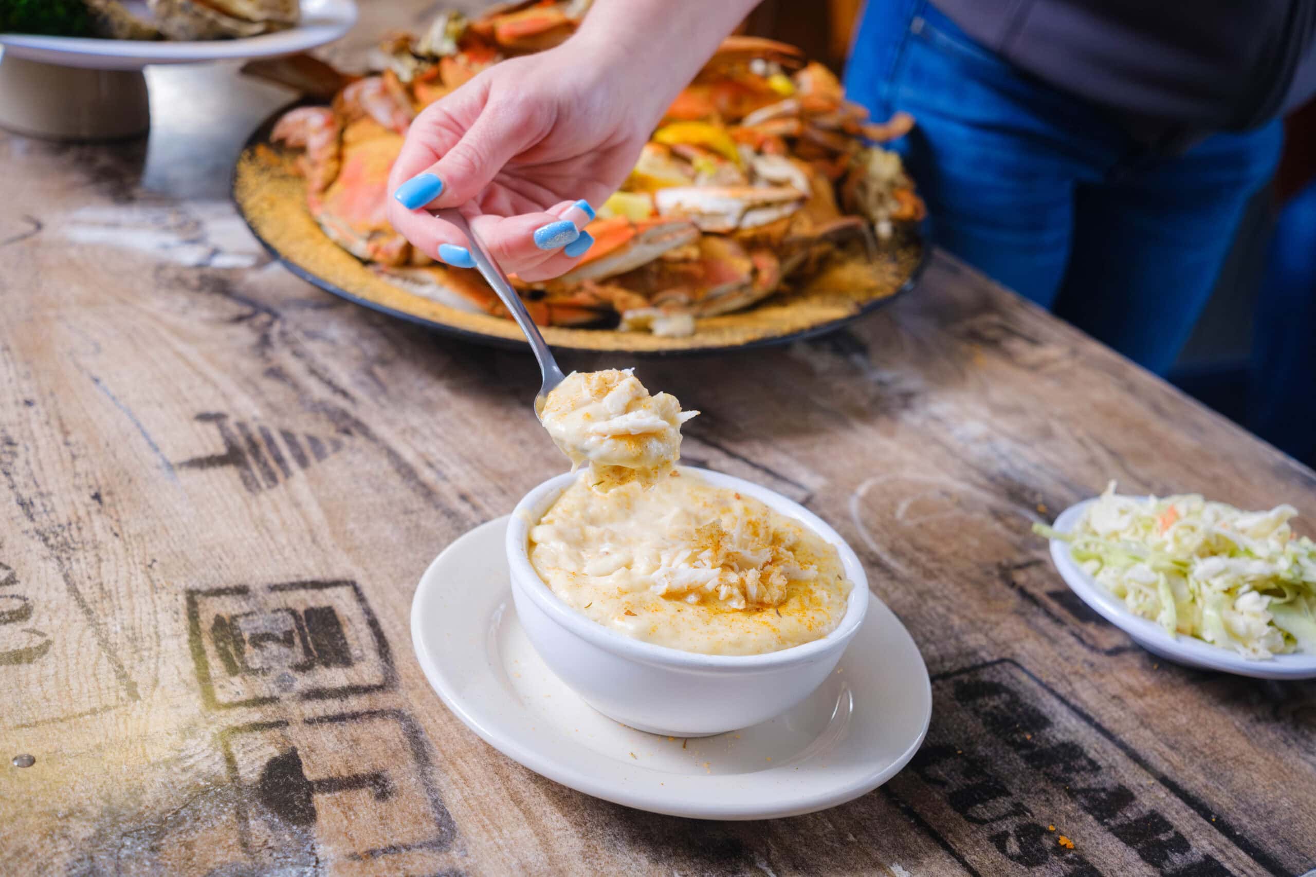 A hand holding a spoonful of creamy she crab soup above a bowl on a wooden table, with a platter of crab in the background and coleslaw on the side at Margie and Ray's Crabhouse & Restaurant.