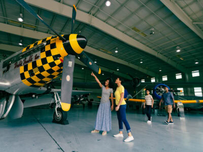 Mother and daughter admire a vintage aircraft with a checkerboard propeller in the Military Aviation Museum hangar.