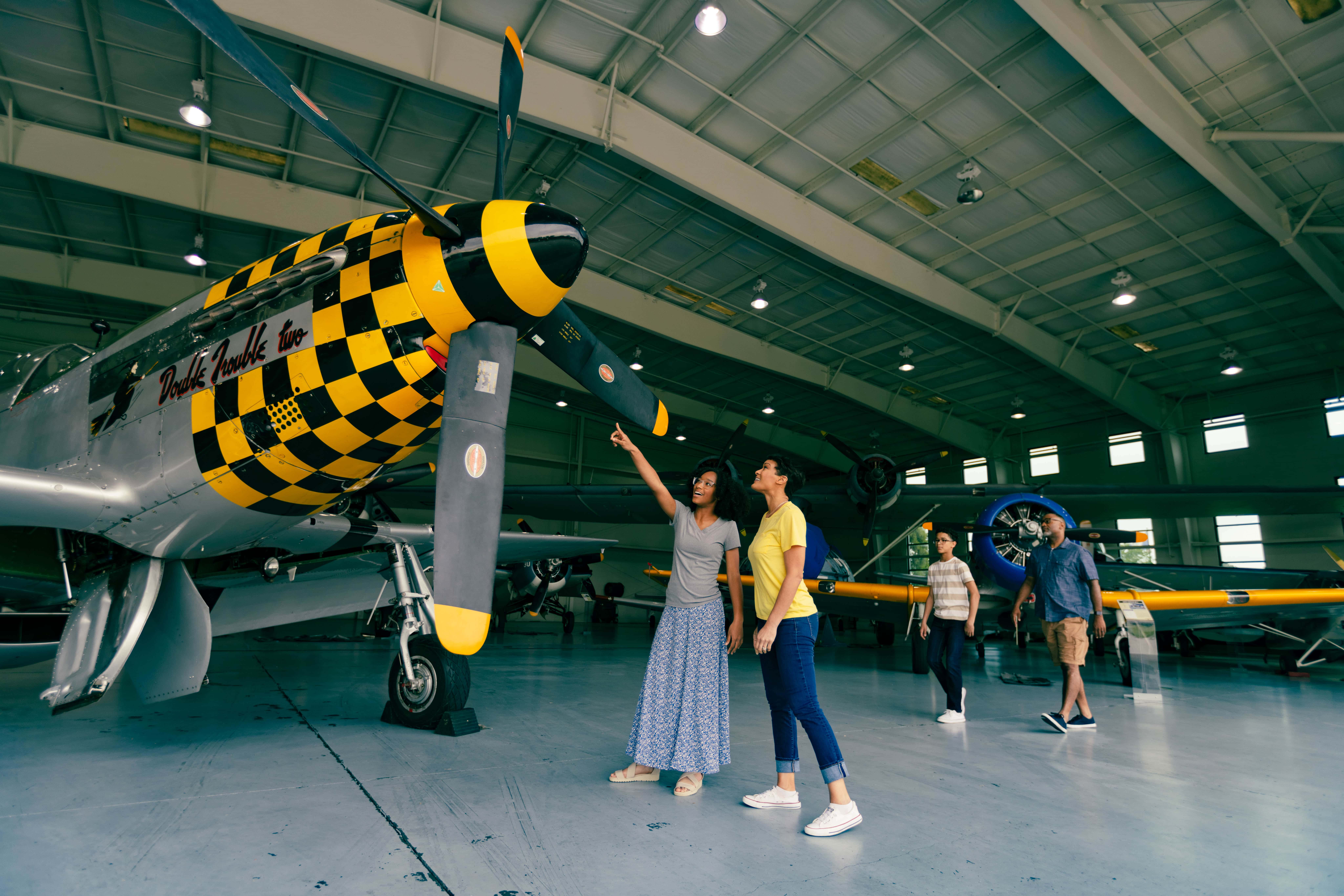 Mother and daughter admire a vintage aircraft with a checkerboard propeller in the Military Aviation Museum hangar.
