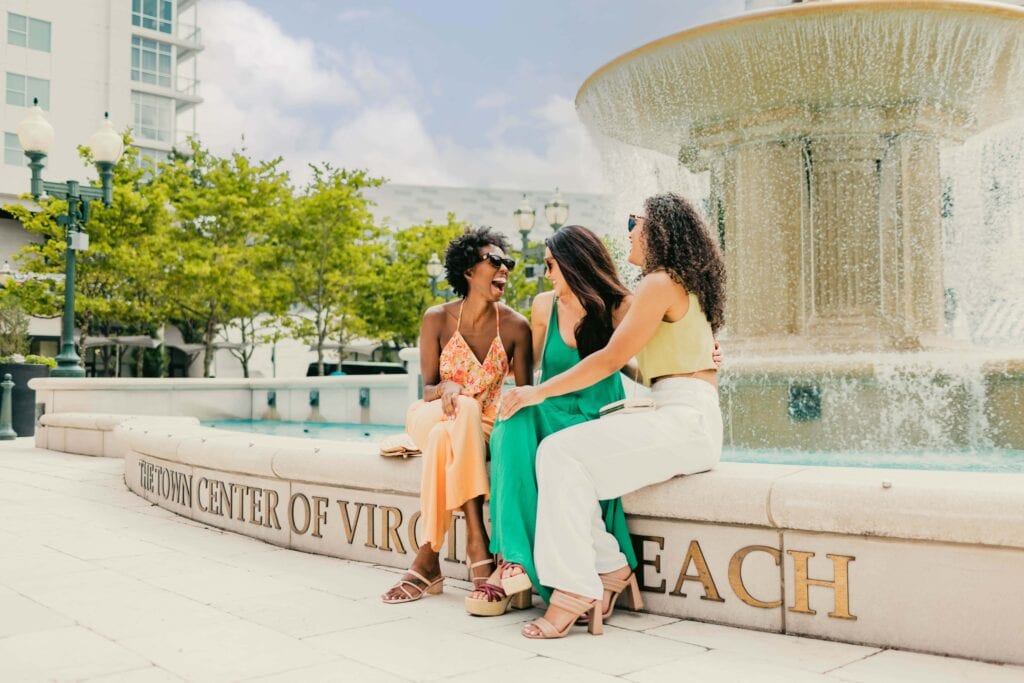 Three women sit on a fountain edge at Town Center of Virginia Beach, laughing and enjoying a sunny day together.