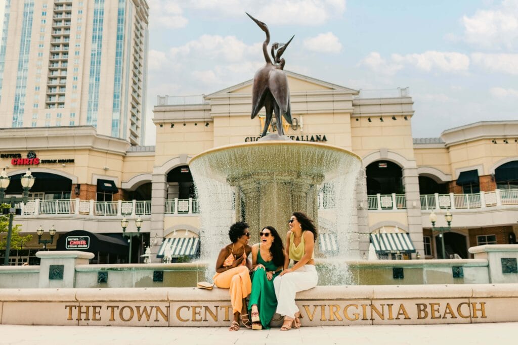 Friends enjoy a sunny day by a fountain at Town Center in Virginia Beach, showcasing vibrant fashion and laughter.