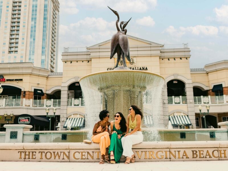 Friends enjoy a sunny day by a fountain at Town Center in Virginia Beach, showcasing vibrant fashion and laughter.
