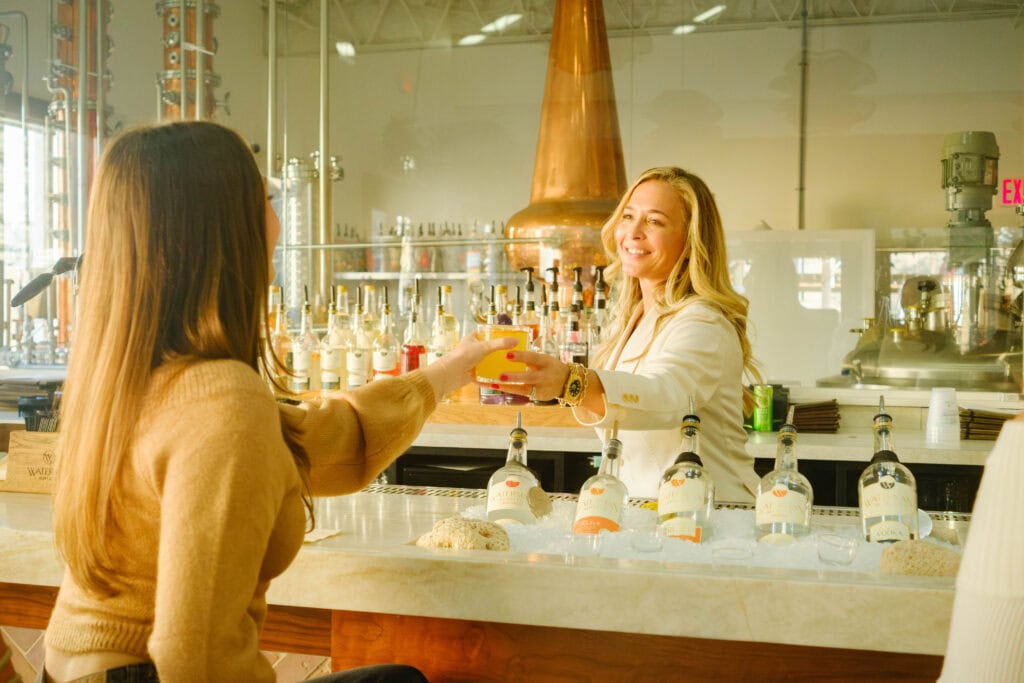 A bartender in a white blazer happily serves a colorful drink to a customer at a stylish bar with liquor bottles displayed & distillery in the background at Mermaid Winery.