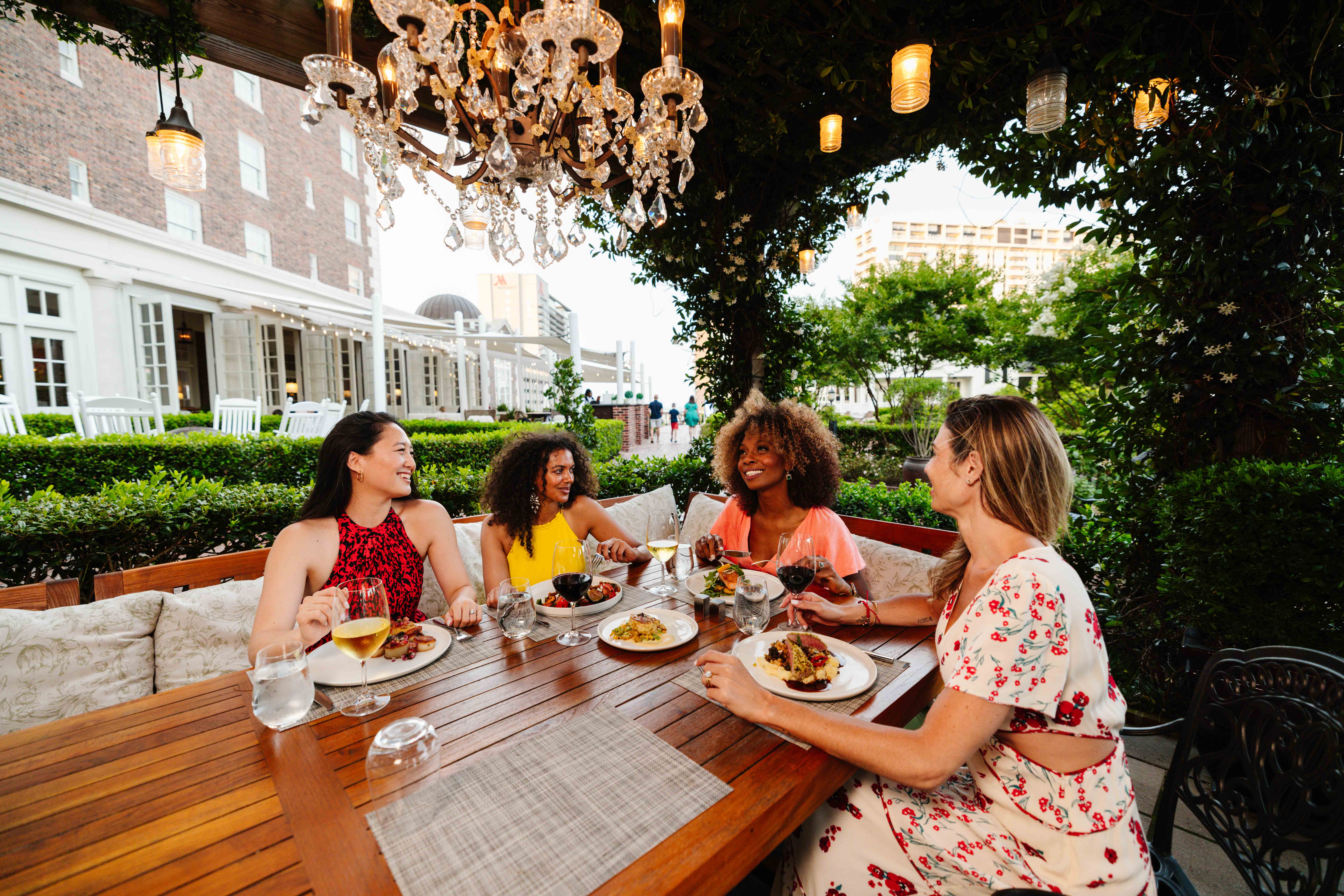 A group of four women enjoying a meal outdoors under a chandelier, with plates of food and drinks on a wooden table amid greenery.