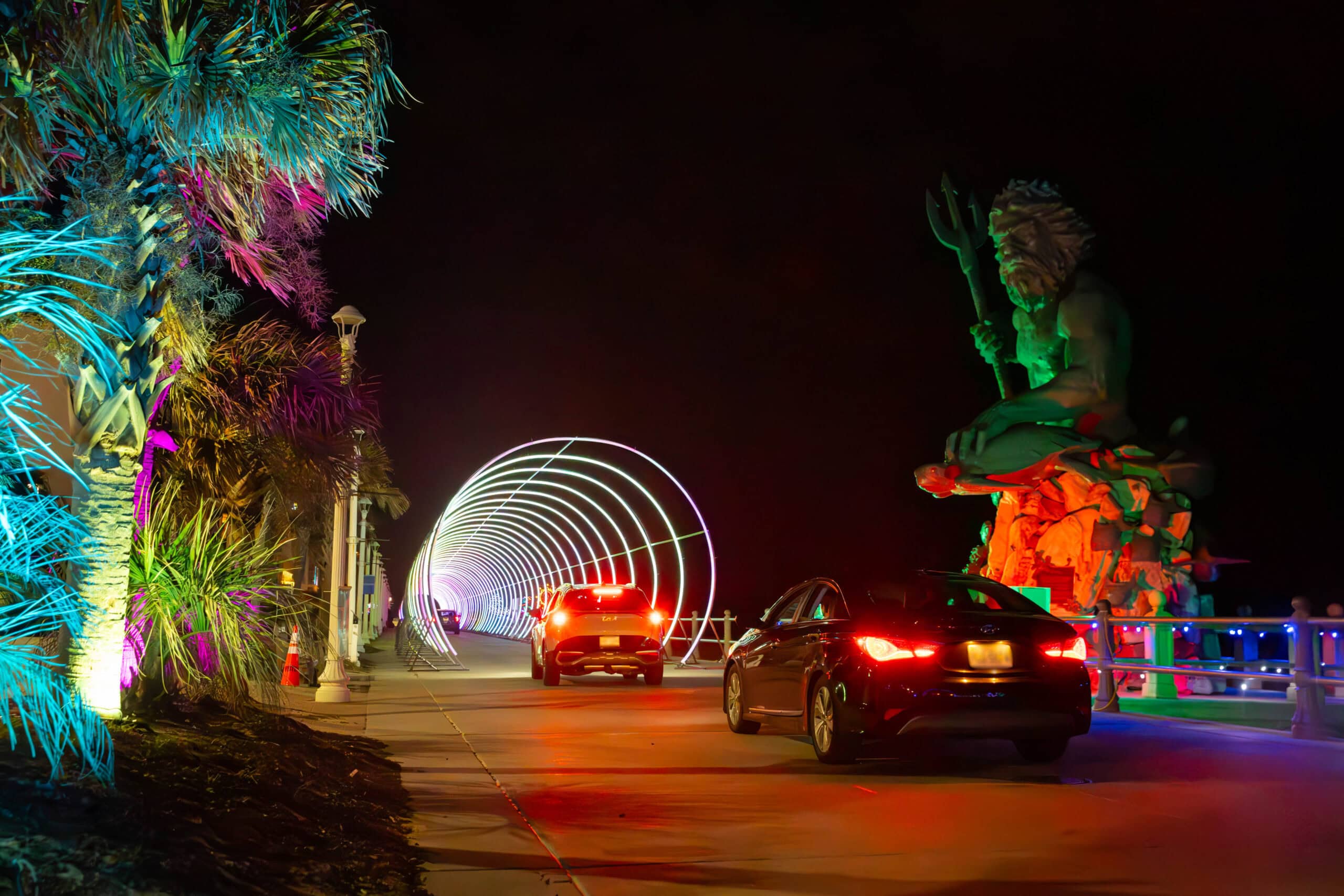 A vibrant nighttime scene with colorful lights illuminating palm trees, cars driving through a glowing arch, and a large statue of King Neptune by the Virginia Beach Boardwalk.