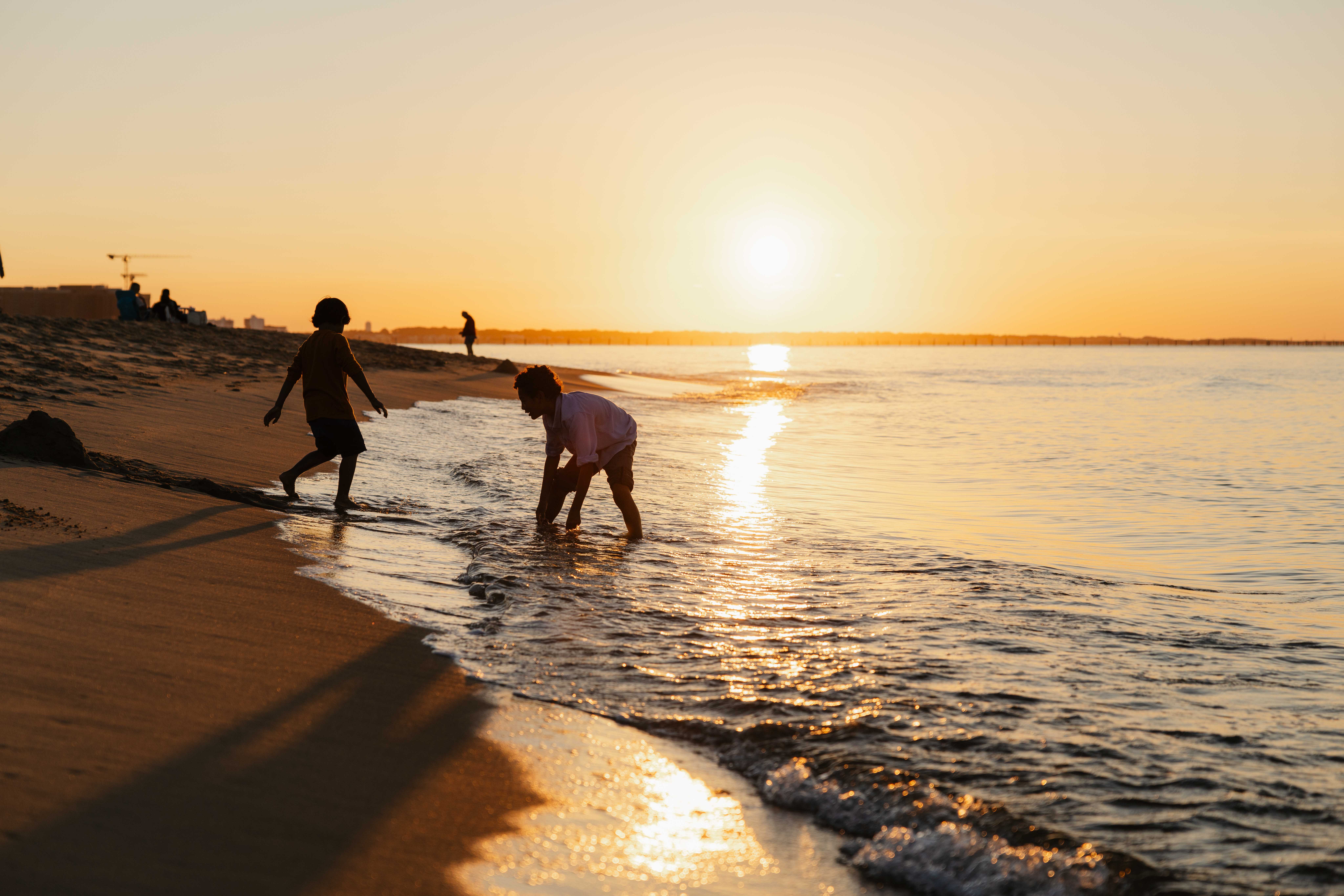 Children playing at sunset on Chesapeake Bay beach, with one child touching the water and the golden sun reflecting on the sea at First Landing State Park.