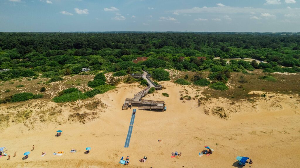 Aerial view of First Landing State Park beach with umbrellas and people, featuring a wooden boardwalk winding through lush green vegetation.