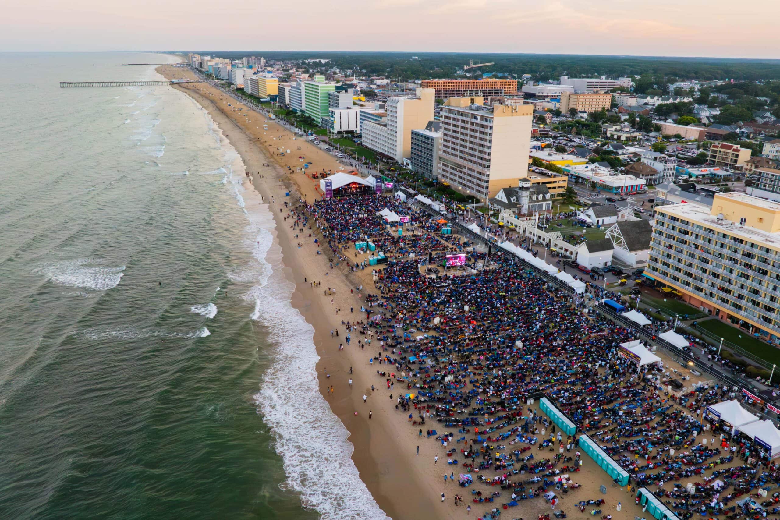 Aerial view of a crowded beach event with numerous people gathered on the sand, coastal buildings, and the ocean in the background.