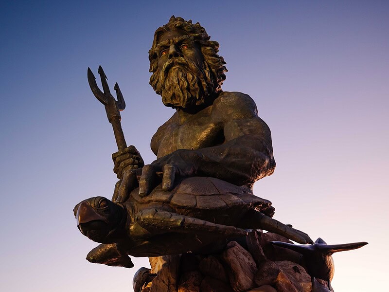 A close up view of the King Neptune Statue holding a trident and a turtle with a purple sunrise sky behind it in Virginia Beach.