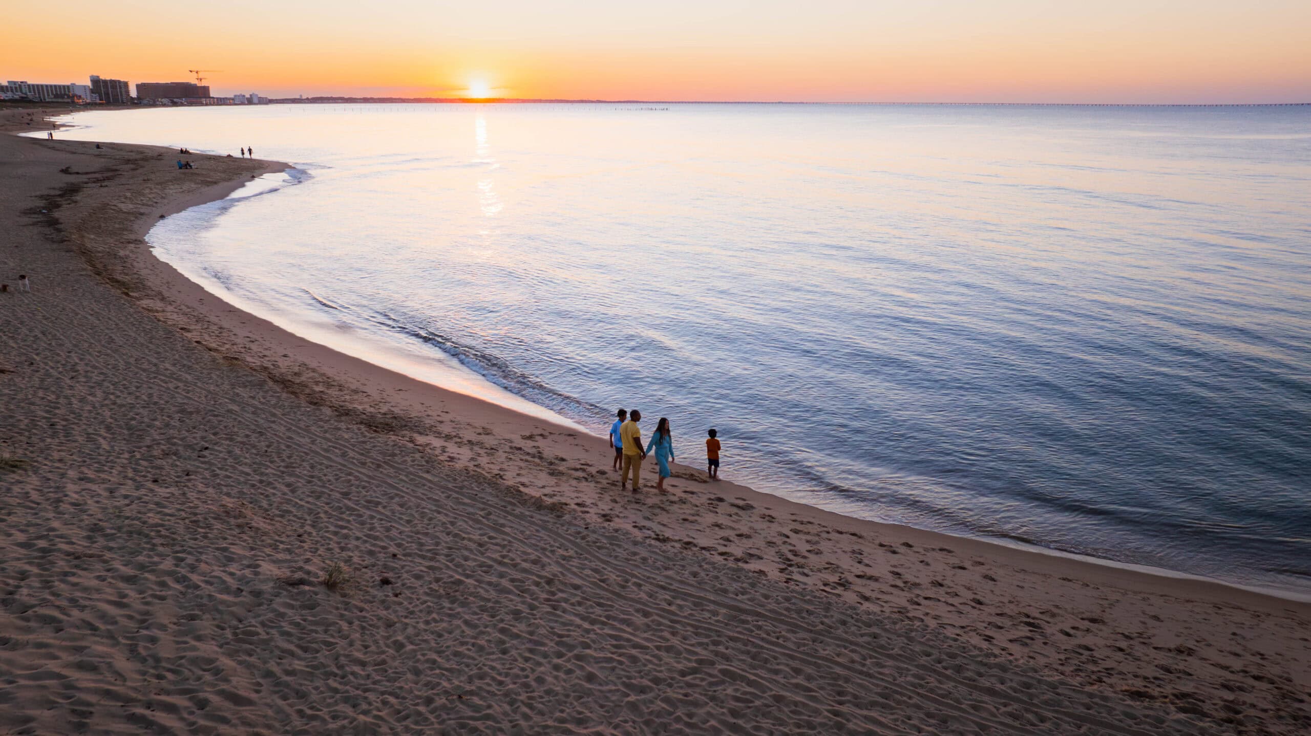 A family walks along a sandy beach at sunset at First Landing State Park, with calm waters reflecting the colorful sky.