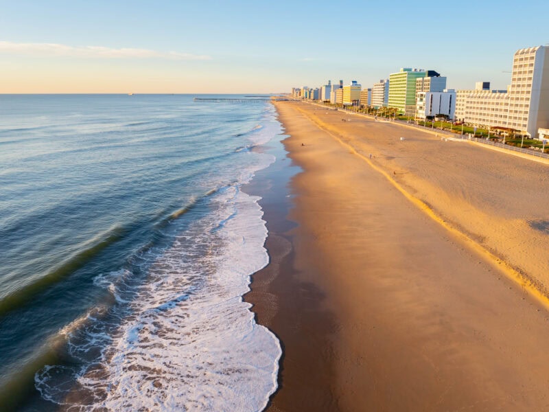 Aerial view of the Virginia Beach Oceanfront with gentle waves lapping at the sandy shore, lined by modern beachfront buildings under a clear sky.