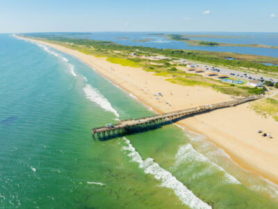 Aerial view of a sandy beach with Little Island pier extending into the turquoise ocean, surrounded by green vegetation and parking areas at Sandbridge Beach.
