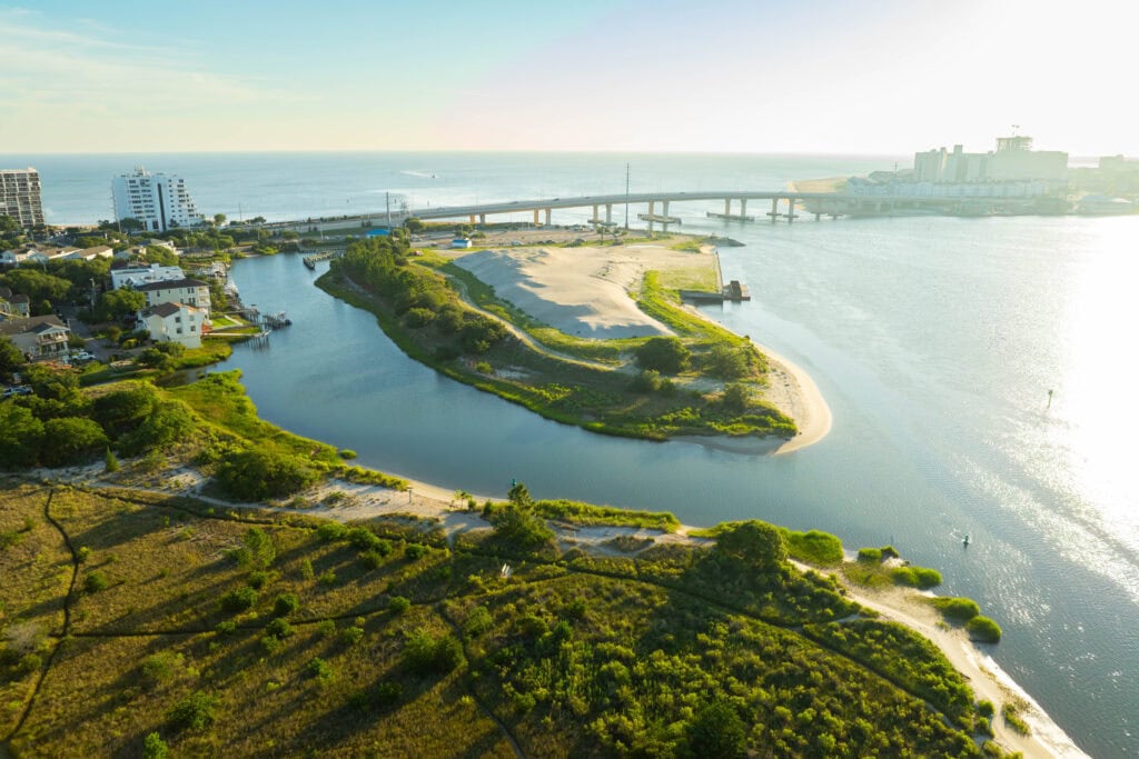 Aerial view of a coastal landscape of Chesapeake Bay featuring a river, sandy banks, greenery, and buildings near the ocean, with Lesner bridge in the background.