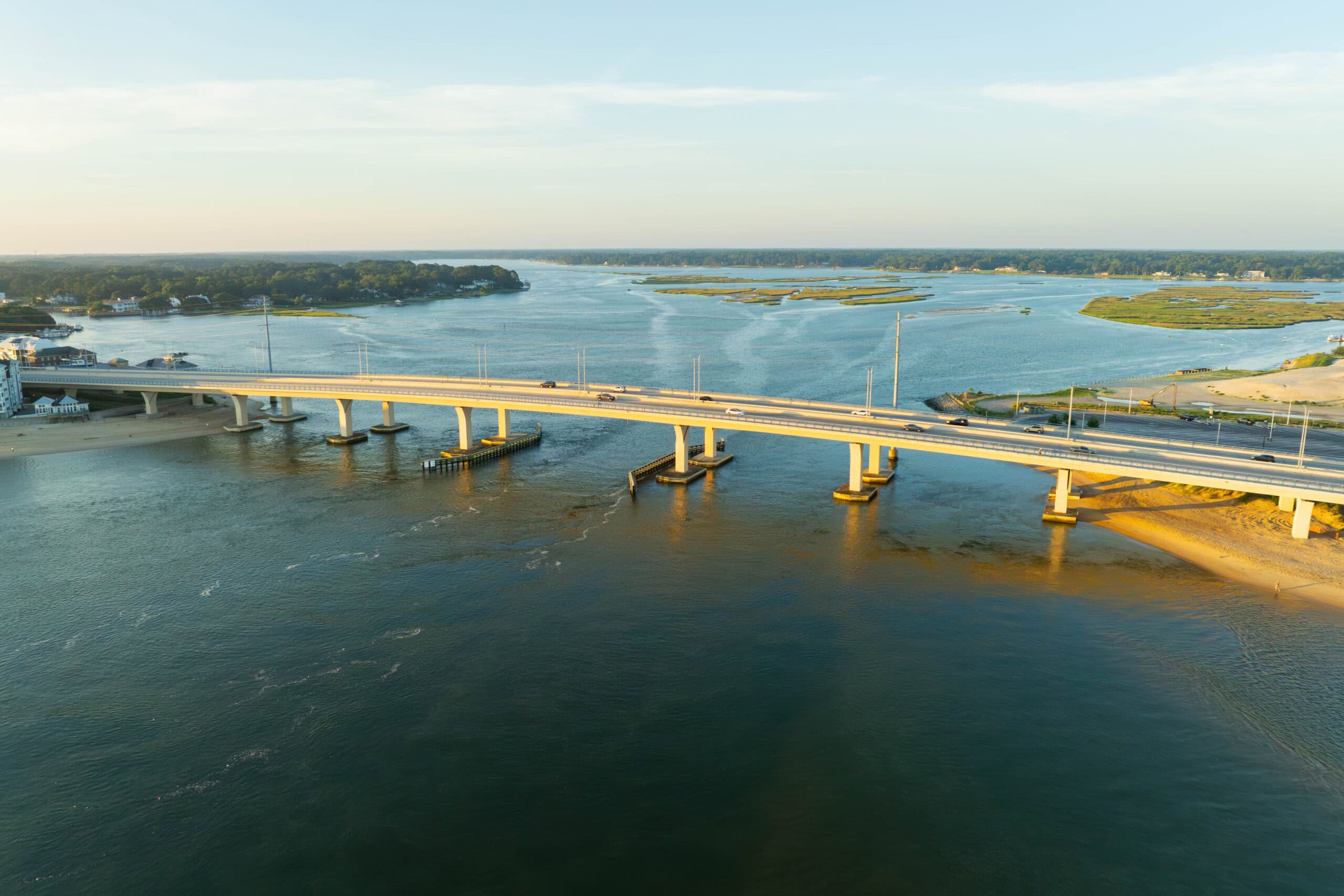 Aerial view of the Lesner bridge spanning the Lynnhaven River, surrounded by lush greenery and sandy shores under a clear sky in Virginia Beach.