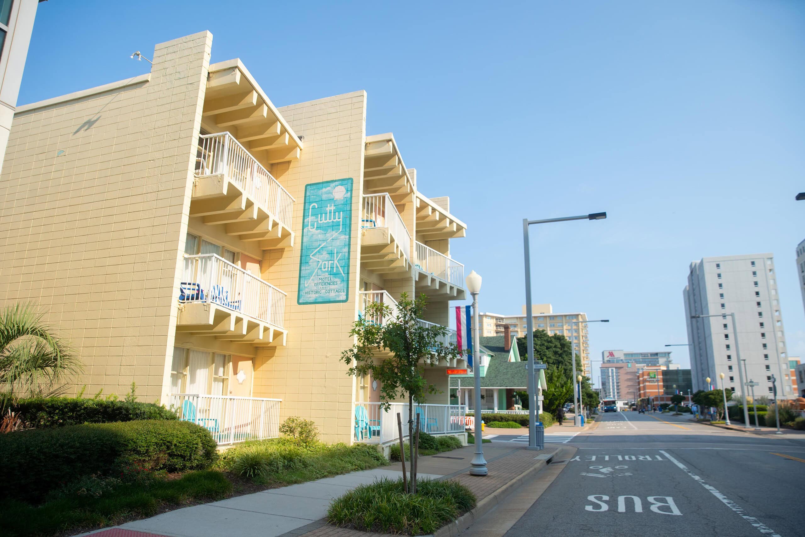 A view of a coastal hotel & cottage named "Cutty Sark," featuring beige buildings with balconies, blue signage, and a street lined with trees and modern buildings in the background at Virginia Beach Oceanfront.